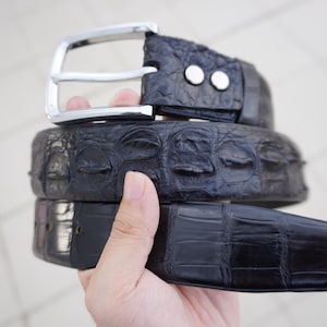 Op de afbeelding: Een zwarte alligatorleren riem met een zilveren gesp. De riem is dubbelgevouwen en wordt in een hand vastgehouden.