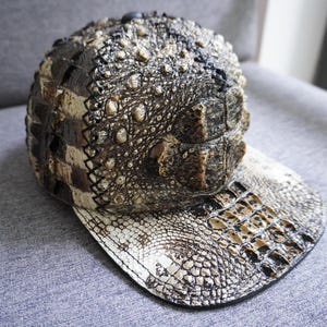 以下が含まれることがあります： 茶色、ベージュ、黒の色合いの、爬虫類のような質感の野球帽。 カーブしたつばと、ワニやアリゲーターの肌を思わせる、詳細なウロコ模様が特徴です。