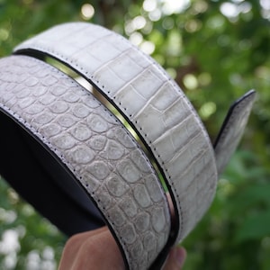 May include: A light gray belt with a textured, crocodile-like pattern. The belt is made of a light gray material with a black interior and a silver-colored buckle. The belt is curved, and the background is blurred green.