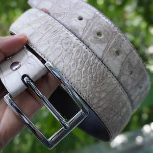 May include: A light gray belt with a textured, reptile-like pattern. The belt has a silver-colored square buckle and a matching silver stud. The belt is made of a durable material and is suitable for everyday wear.
