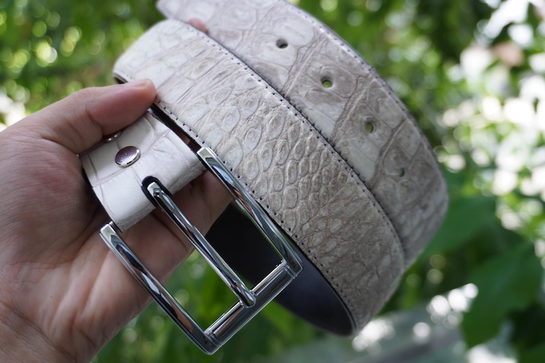 May include: A light gray belt with a textured, reptile-like pattern. The belt has a silver-colored square buckle and a matching silver stud. The belt is made of a durable material and is suitable for everyday wear.