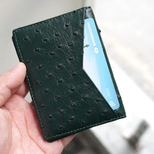 May include: Dark green leather card holder with a textured surface. A light blue debit card is partially visible in the front pocket. The card holder is held in a hand.