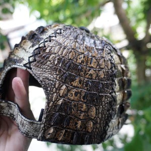 K&ouml;nnte beinhalten: Baseballcap mit einem strukturierten, braunen und beigefarbenen Krokodilhautmuster. Die Kappe hat einen gebogenen Schirm, eine goldfarbene Metallschnalle und schwarze Nahtdetails. Ein einzigartiges Accessoire.