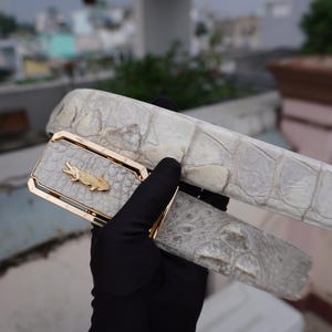 May include: A white leather belt with a gold buckle featuring an alligator design. The belt is made of a textured material and has a unique, embossed pattern.