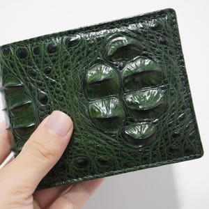Puede incluir: Una cartera de cuero verde oscuro con textura de piel de cocodrilo. La cartera es rectangular y tiene un diseño plegable. La superficie tiene un patrón irregular en forma de escamas. La cartera se sostiene en una mano sobre un fondo blanco.