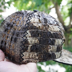 K&ouml;nnte beinhalten: Eine Kappe aus strukturiertem Material mit gebogenem Schirm und verstellbarem Riemen. Die Oberfl&auml;che der Kappe zeigt eine Mischung aus Braun-, Beige- und Schwarzt&ouml;nen mit sichtbaren N&auml;hten. Die Kappe ist im klassischen Baseballkappen-Stil gestaltet.