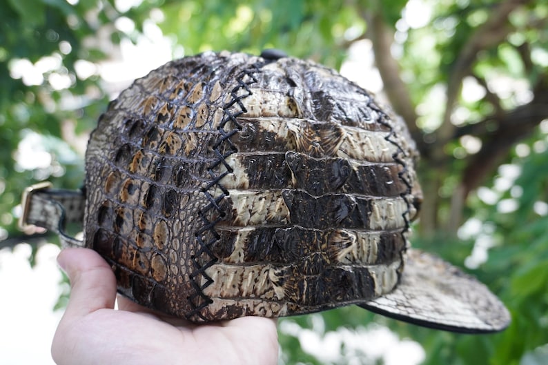K&ouml;nnte beinhalten: Eine Kappe aus strukturiertem Material mit gebogenem Schirm und verstellbarem Riemen. Die Oberfl&auml;che der Kappe zeigt eine Mischung aus Braun-, Beige- und Schwarzt&ouml;nen mit sichtbaren N&auml;hten. Die Kappe ist im klassischen Baseballkappen-Stil gestaltet.