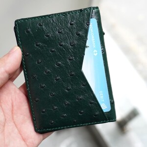 May include: A dark green leather wallet with a textured surface and a card slot. The wallet is held in a hand, and a blue credit card is partially visible in the slot. The wallet's dimensions are approximately 4 inches by 3 inches.