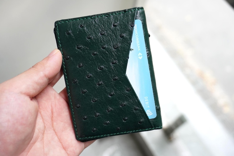May include: A dark green leather wallet with a textured surface and a card slot. The wallet is held in a hand, and a blue credit card is partially visible in the slot. The wallet's dimensions are approximately 4 inches by 3 inches.