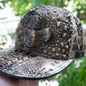 K&ouml;nnte beinhalten: Baseballkappe mit einem strukturierten Design, das Krokodilleder &auml;hnelt. Die Kappe hat einen gebogenen Schirm und ein detailliertes Muster aus Schuppen und Unebenheiten in Braun-, Beige- und Schwarzt&ouml;nen. Die Kappe wird in einer Hand gehalten.