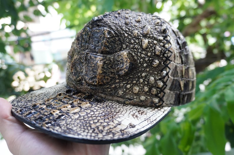 K&ouml;nnte beinhalten: Baseballkappe mit einem strukturierten Design, das Krokodilleder &auml;hnelt. Die Kappe hat einen gebogenen Schirm und ein detailliertes Muster aus Schuppen und Unebenheiten in Braun-, Beige- und Schwarzt&ouml;nen. Die Kappe wird in einer Hand gehalten.