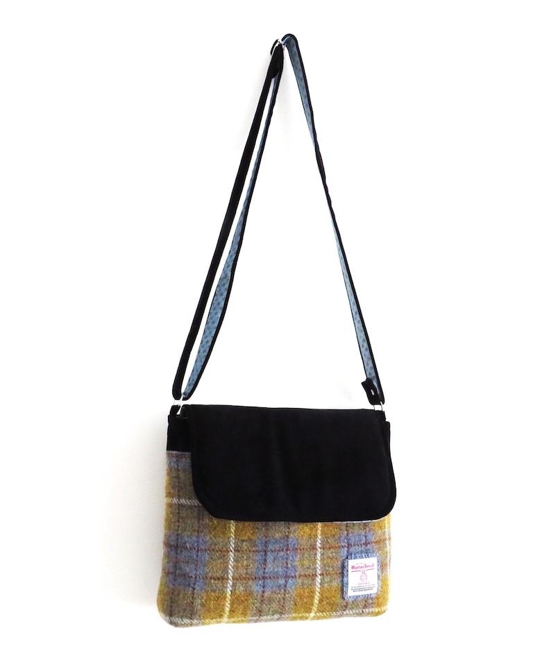 May include: A shoulder bag with a black flap and strap, and a plaid pattern in yellow, blue, and brown. The bag is hanging against a white background. The strap is black with a blue patterned lining.