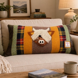 May include: A rectangular throw pillow featuring a central applique of a white Highland cow head on a mustard yellow and brown herringbone background. The sides are accented with green and multi-colored plaid fabric panels and a small rectangular label.
