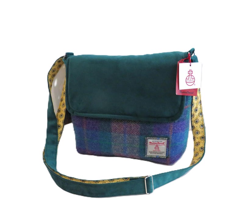 May include: A teal and plaid Harris Tweed shoulder bag with a flap closure. The bag features a teal strap with a yellow paisley pattern on the underside. A tag with a logo hangs from the top.