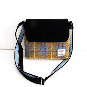 May include: A shoulder bag with a black flap and strap. The bag's body features a yellow, blue, and red plaid pattern. The strap is black with a blue patterned trim. A small label is sewn on the front.