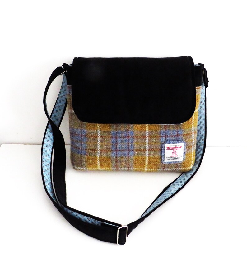 May include: A shoulder bag with a black flap and strap. The bag's body features a yellow, blue, and red plaid pattern. The strap is black with a blue patterned trim. A small label is sewn on the front.