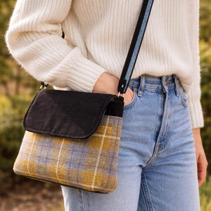 May include: A crossbody bag with a black flap and a plaid pattern in yellow, blue, and white. The bag has a black and blue strap. The bag is worn over a white sweater and blue jeans.