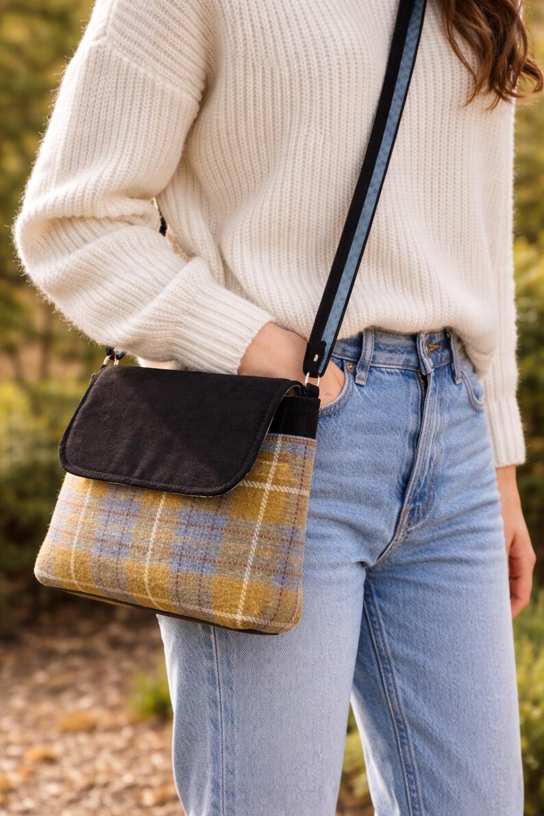 May include: A crossbody bag with a black flap and a plaid pattern in yellow, blue, and white. The bag has a black and blue strap. The bag is worn over a white sweater and blue jeans.