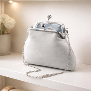 May include: A light gray, fabric clutch purse with a silver metal clasp and chain strap. The interior lining features a blue floral pattern. The purse is sitting on a white shelf.