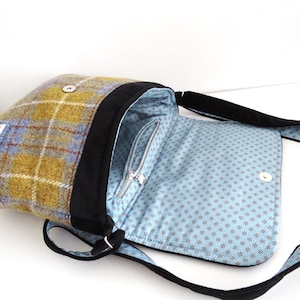 May include: A shoulder bag with a plaid exterior in shades of yellow, blue, and brown. The bag has a black trim and strap, with a light blue interior patterned with small, dark red flowers. The bag is open, revealing a zippered pocket.