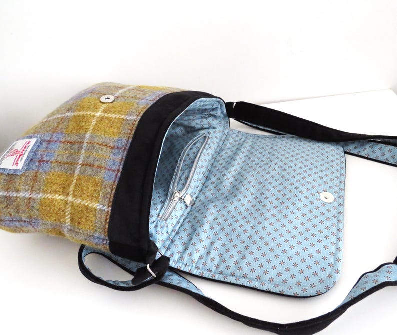 May include: A shoulder bag with a plaid exterior in shades of yellow, blue, and brown. The bag has a black trim and strap, with a light blue interior patterned with small, dark red flowers. The bag is open, revealing a zippered pocket.