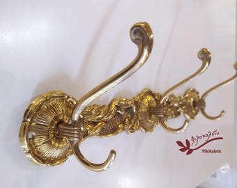 May include: A gold-toned metal wall-mounted coat rack featuring three ornate hooks with curved ends. The base plates are circular with a fluted, shell-like design, and the connecting bar displays intricate relief patterns. Text reads Filokalein.