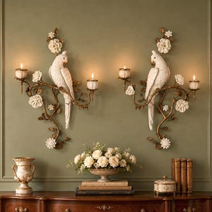 May include: A pair of wall-mounted candle sconces featuring white cockatoo figures perched on gold-toned branches adorned with white floral accents. Each sconce holds two small white candle cups with lit candles, creating a symmetrical display.