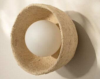 May include: A beige ceramic wall sconce with a round, white frosted glass light bulb. The sconce has a rough, textured surface.