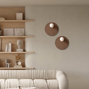 May include: A white sofa with a tufted design sits in a living room with a wooden shelf unit, a coffee table with a marble top, and a potted plant. Two round wall sconces with woven shades are on the wall.