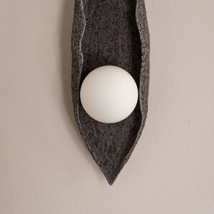 Puede incluir: Una lámpara de pared de cerámica gris con una luminaria esférica blanca en el centro. La lámpara tiene forma de hoja y una superficie texturizada.