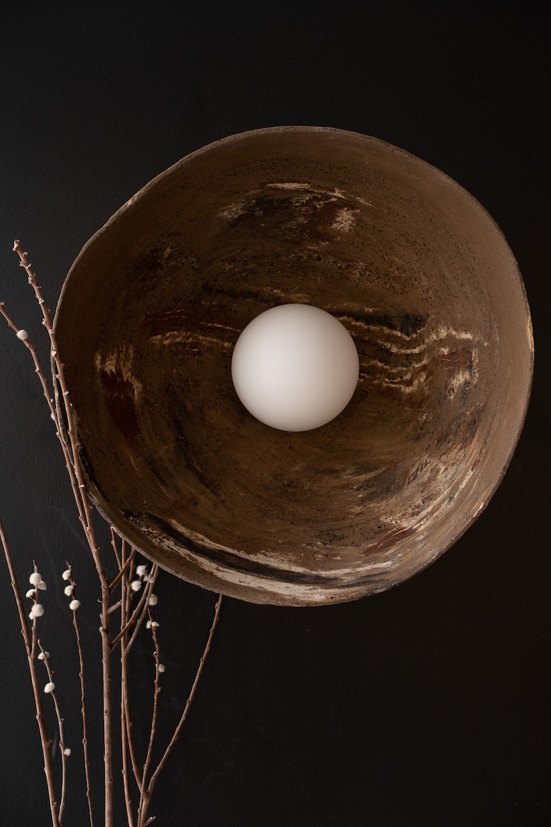 May include: A white orb sits in the center of a large, rustic brown bowl with a textured surface. The bowl is set against a black background.