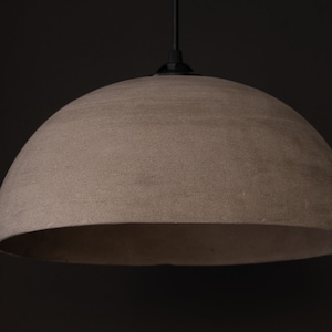 May include: A dome-shaped pendant light fitting with a textured, neutral-toned lampshade. The light hangs from a black cord and canopy against a dark background. The lampshade has a semi-circular shape.