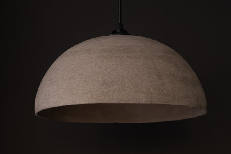 May include: A dome-shaped pendant light fitting with a textured, neutral-toned lampshade. The light hangs from a black cord and canopy against a dark background. The lampshade has a semi-circular shape.
