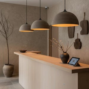 May include: Interior view of a contemporary space with three pendant lights with grey dome shades suspended above a light wood counter. A vase with branches and a tablet are on the counter. The walls are a textured beige.