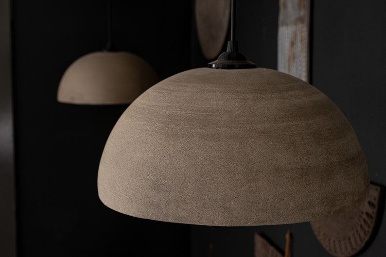 May include: Two pendant lights with textured, dome-shaped shades in a neutral beige colour. The lights hang from black cords against a dark background. The lampshades have a speckled appearance.