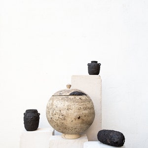Op de afbeelding: Een witte keramische pot met deksel en een ruwe textuur staat op een wit voetstuk. Twee kleinere zwarte keramische potten staan op witte voetstukken achter de grotere pot. Een zwarte ovale keramische vorm staat op een wit voetstuk voor de grotere pot.