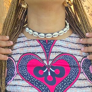 以下が含まれることがあります： ハート柄のピンクとブルーのクロップトップで、長袖です。トップは白い背景に模様のある生地で作られています。モデルは白い貝殻のネックレスを付けてトップを着ています。