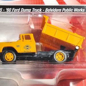 May include: A yellow '60 Ford Dump Truck model from Belvedere Public Works. The truck has a black chassis and yellow dump bed. The text on the truck reads "3055" and "Belvidere". The model is packaged in a clear plastic case.