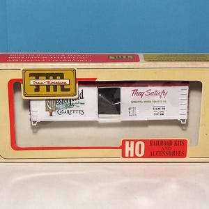 May include: A white box with a red and gold border. The box has a picture of a train car with the words "Chesterfield Cigarettes" and "They Satisfy" on it. The box also has the words "HO Railroad Kits and Accessories" on it.