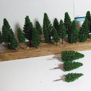 May include: Miniature green trees with brown trunks are arranged on a wooden base. Several additional trees are placed separately. These model trees are likely for a diorama or model train landscape.