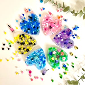 Puede incluir: Cinco artesanías de resina en forma de corazón llenas de cuentas de colores. Cada corazón presenta un tema diferente: azul con cuentas blancas y negras, amarillo con caritas sonrientes negras, morado con cuentas blancas y plateadas, rosa con cuentas blancas y rosas, y verde con flores blancas y rosas.