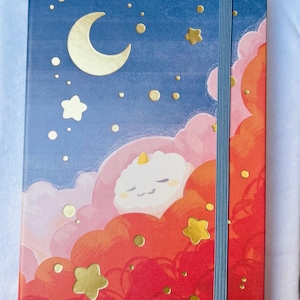 Può includere: Un taccuino rettangolare con un design celeste. La copertina presenta un cielo blu con una luna a mezzaluna dorata, stelle e nuvole nelle tonalità del rosa, arancione e rosso. Un elastico blu assicura il taccuino.