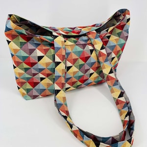 Puede incluir: Una bolsa de mano de lona con estampado geométrico de colores y dos asas largas. La bolsa está hecha de un tejido tejido con una variedad de colores, incluyendo rojo, amarillo, azul, verde y negro.
