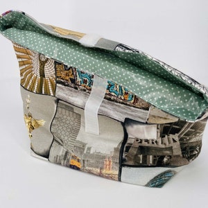 Puede incluir: Una bolsa de tela gris y blanca con un diseño de patchwork que presenta imágenes de edificios y paisajes urbanos. La bolsa tiene un forro de lunares verdes y un cierre de velcro blanco.