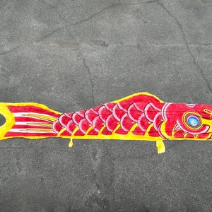 Puede incluir: Una colorida manga de viento con forma de carpa, de color rojo con detalles amarillos y blancos. El pez tiene un patrón de escamas detallado y una cola amarilla. Diseñado para colgar al aire libre.