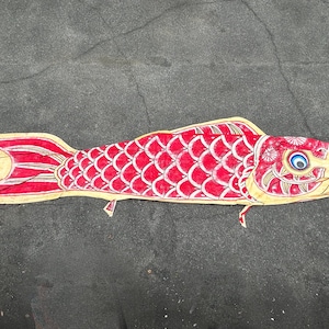 May include: A red and white carp-shaped windsock, or koinobori, with a yellow outline. The fish has detailed scales and fins, and a blue and white eye. The windsock is lying on a dark grey surface.
