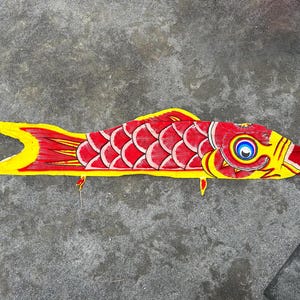Puede incluir: Cometa artesanal con forma de pez. El cuerpo es rojo con escamas blancas, aletas y cola amarillas. La cabeza es amarilla con un ojo azul y blanco. Descansa sobre una superficie gris.