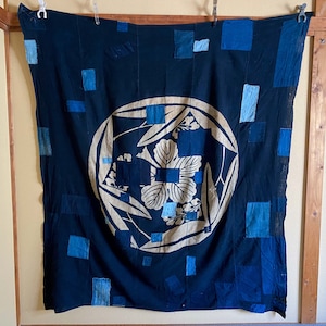 May include: A dark blue fabric wall hanging with a white floral design in the centre. The fabric is made up of many small squares of blue fabric sewn together.