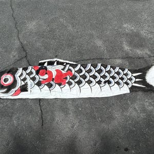 Puede incluir: Una gran manga de viento con forma de carpa blanca con detalles en negro, rojo y gris. El pez tiene un patrón de escamas detallado, detalles en rojo y una cara sonriente. La manga de viento está sobre una superficie gris.