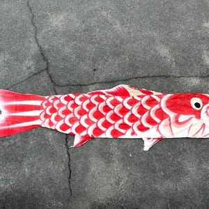 May include: A red and white carp streamer, a traditional Japanese decoration. The carp-shaped windsock lies on a grey surface. The fish has a red body with white scale patterns and a white tail.